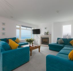 A living room with blue sofas and yellow cushions a wooden table with flowers and a fireplace with a TV on the wall at Moonbeams in Carbis Bay