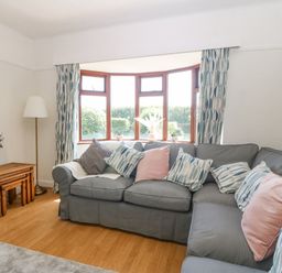 A living room with a gray sectional sofa and patterned cushions near a bay window with blue patterned curtains and nested wooden tables at Awelfryn in Elim near Llanddeusant