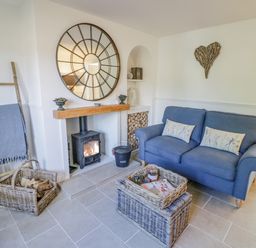 A living room with a blue sofa a wood burning stove and wicker baskets at Blossom Cottage in Broadway