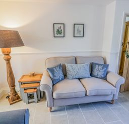 A living room with a sofa with three cushions a wooden floor lamp a set of nesting tables and a wooden door at Blossom Cottage in Broadway