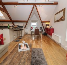 An open plan living and dining area with wooden beams a wooden floor a kitchen island with bar stools a dining table with chairs and a sofa with red cushions at Apartment 2 Gorwel Glas in Llanbedrog