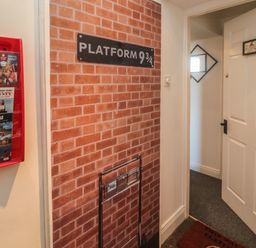 An indoor hallway with a brick wall decal and platform 9 3/4 sign next to a white door and a red brochure holder with maps at The Wizards Loft in Alnwick