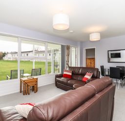 A living room with brown leather sofas red and white cushions a wooden nested table large windows and a dining table with black chairs at Marsh Bungalow in Lowestoft