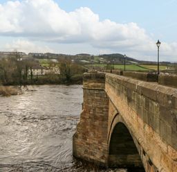 A stone bridge over a flowing river with lampposts along the bridge and fields in the background at Bramwell House in Corbridge