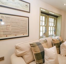A sitting area with a beige sofa and patterned cushions under a window with two vases and framed documents on the wall at Bramwell House in Corbridge