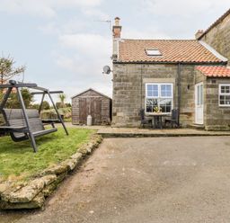 An outdoor area with a stone cottage a wooden swing on grass a stone shed and a table with two chairs at Hawthorn Dale Cottage Whitby