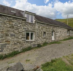 Stone house with multiple windows and a white door on a gravel path with green hills and cloudy sky at Pennant Tigi Uchaf in Dinas Mawddwy