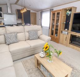 A living room with a beige sofa coffee table with sunflowers and a vase wooden cabinet television and kitchen in the background at Chantry Lodge Hendra Croft near Goonhavern