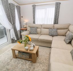 A living room with a beige sectional sofa a wooden coffee table with sunflowers and teapot set near windows with curtains at Chantry Lodge Hendra Croft near Goonhavern