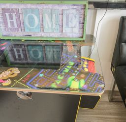 A vintage arcade game table with pac-man and donkey kong graphics with a framed home sign on top and a black leather chair next to it at Wyvill House in Cayton