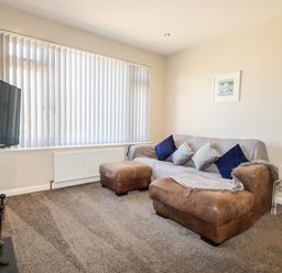 A living room with a brown leather sofa and ottoman a wall-mounted television and vertical blinds on the window at Seawynds in Crantock