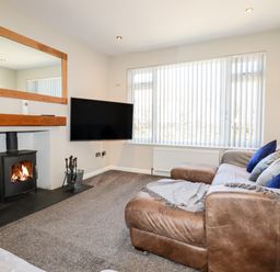 A living room with a wood stove fireplace a TV on the wall a leather sofa with cushions and a large window with vertical blinds at Seawynds in Crantock