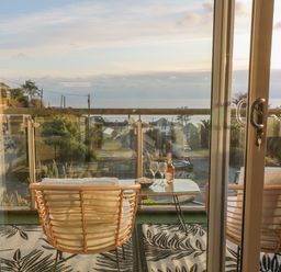 A balcony with two cushioned chairs a glass table with a bottle and glasses overlooking houses and the sea at A Stone's Throw in Downderry