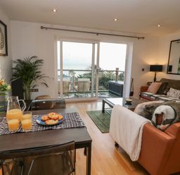 A living room with a dining table set with juice and pastries and a seating area with sofas and a coffee table at A Stone's Throw in Downderry
