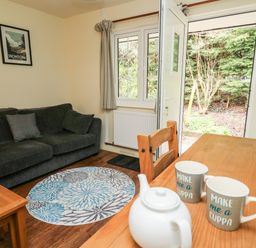 A living room with a dark sofa a round patterned rug a wooden table with teapot and mugs and an open door to a garden at Honeysuckle Cottage in Crossgates