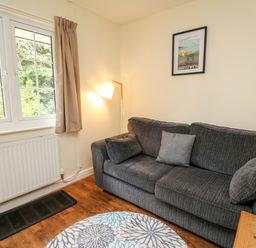 A sitting area with a grey sofa, cushions, a floor lamp, a window with beige curtains, a radiator, a round patterned rug and a framed picture on the wall at Primrose Cottage in Crossgates