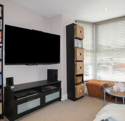 A living room with a wall-mounted television a media console bookshelf with DVDs a tall shelving unit with wicker baskets an ottoman by windows with blinds and a small table with a tea set at Alltwen in Borth-y-Gest