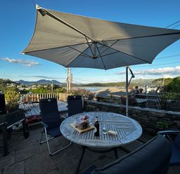 An outdoor patio with a wooden table under a large umbrella surrounded by chairs and a barbecue grill at Alltwen in Borth-y-Gest