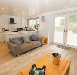 An open living area with a brown sofa, wooden coffee table, and a kitchen with white cabinets at Bryn Eglwys Bach in Penmynydd near Llanfairpwllgwyngyll