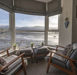 A sitting area with two armchairs, two ottomans, small plants on the window sill, and a view of the water and hills at Harbour View - Flat 2 in Barmouth