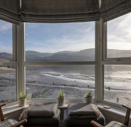A bay window with stools blankets small plants and chairs overlooking a tidal shore with hills in the distance at Harbour View - Flat 2 in Barmouth