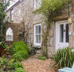 An outdoor stone wall with a white door and window surrounded by plants and a mirror on a lattice with a light blue bench at Rose Cottage in Lower Bostraze near St Just