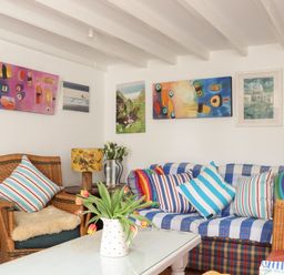 A sitting area with wicker chairs a sofa with striped cushions a white coffee table with flowers and paintings on the wall at Rose Cottage in Lower Bostraze near St Just