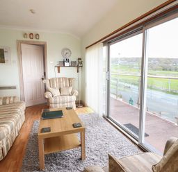 A sitting room with striped sofas a wooden coffee table a wall clock and large sliding glass doors overlooking a road and fields at Morlyn Guest House Apartment in Llandanwg near Harlech