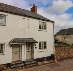 A white cottage with two green doors and windows next to a stone wall at Bodkin Cottage in Dunster