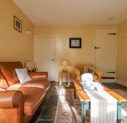 A living room with a brown leather sofa an armchair a wooden coffee table with a table runner and tea set a wall clock and doors at Bodkin Cottage in Dunster