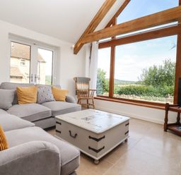 A living room with a gray sofa yellow pillows a white coffee table large window with wooden frame and a wooden chair at The Nook in Jack's Green near Painswick