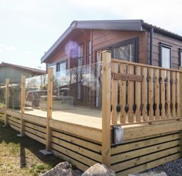 An exterior view of a wooden lodge with glass panel railings and outdoor seating at Curlew Lodge in Carnforth