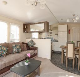 A living room with a sofa and cushions a coffee table with a plant a dining table with chairs and a kitchen area at Audrey's Lodge Foxburrow Hang in Fritton