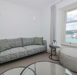 A sitting area with a grey patterned sofa two cushions round ottoman side table with binoculars and magazine window with a view at Upper Tremorvah in Dartmouth