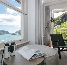 A room with a chair lamp and table with binoculars and magazine near windows overlooking water and trees at Upper Tremorvah in Dartmouth