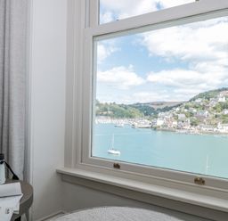 A window with a view of boats on water and houses on hills with curtains and binoculars on a table at Upper Tremorvah in Dartmouth
