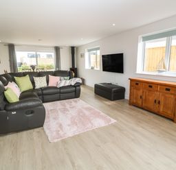 A living room with a black sectional sofa with cushions a wooden sideboard a wall mounted tv a small wood burning stove and large windows at 3 Praa Cove in Praa Sands