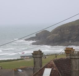 A coastal scene with rooftops and chimneys in the foreground a grassy area with people walking and a flag near the shore and rocky cliffs by the sea at 3 Praa Cove in Praa Sands