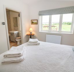 A bedroom with a white bedspread and folded towels on the bed a bedside table with a lamp and a window showing horses outside at Flagstone Cottage in Upper Swell near Stow-On-The-Wold