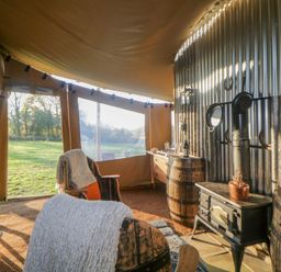 An interior view of a tent with chairs, a stove, wooden barrels, and a window overlooking grass at The Wagon at Burrow Hill in Ottery St Mary