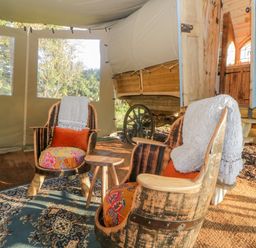 Two wooden chairs with cushions and throws inside a tent next to a wooden wagon at The Wagon at Burrow Hill in Ottery St Mary