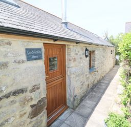 Stone exterior wall with wooden door and small window along a paved path beside green hedges at Godolphin Barn Ashton near Rosudgeon