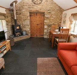 A rustic living room with a wood burning stove dining table orange sofa and television at Godolphin Barn in Ashton near Rosudgeon