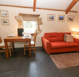 A room with a red sofa a small wooden dining table with two chairs and framed pictures on the wall at Godolphin Barn in Ashton near Rosudgeon