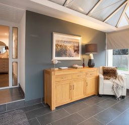 A room with a wooden sideboard, a framed picture above it, a lamp, and a white armchair with a cushion and blanket at Hafod Heli in Rhosneigr