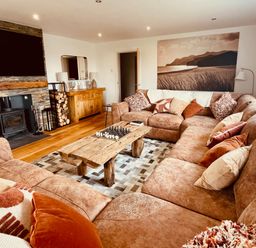 A living room with a large brown sectional sofa and a wooden coffee table with chess pieces a fireplace with a wood-burning stove a flat-screen TV above the fireplace a wooden sideboard with lamps a framed landscape photo on the wall at Hafod Heli in Rhosneigr