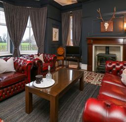A living room with red leather sofas a wooden coffee table a fireplace and windows with curtains at Tirallen in Llangadog