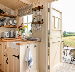 A kitchen corner with shelves of dishes and utensils and an open door leading outside at High Grounds Shepherd's Hut Boylestone near Ashbourne