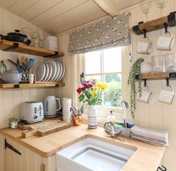 A small kitchen corner with wooden shelves holding plates cups and pans near a window with flowers on the countertop at High Grounds Shepherd's Hut Boylestone near Ashbourne