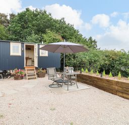 An outdoor patio area with a table and two chairs under an umbrella in front of a black corrugated metal shepherd's hut at High Grounds Shepherd's Hut in Boylestone near Ashbourne